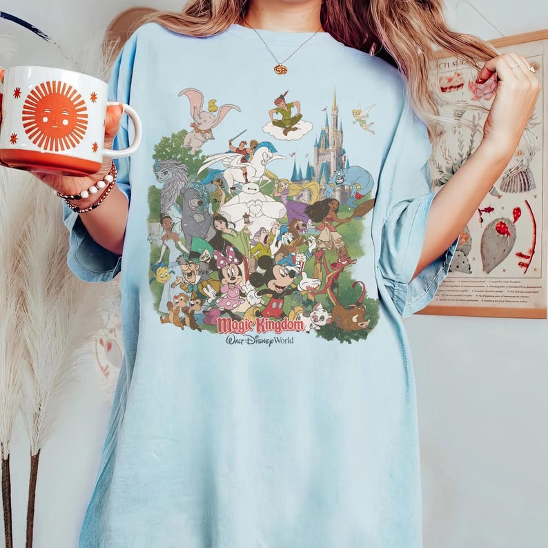 K&ouml;nnte beinhalten: Hellblaues T-Shirt mit einem farbenfrohen Aufdruck von Disney-Figuren, darunter Mickey Mouse und Dumbo. Der Text "Magic Kingdom Walt Disney World" ist darunter gedruckt. Eine wei&szlig;e und orangefarbene Tasse mit einem Sonnendesign wird im Bild gehalten.