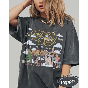 May include: A charcoal grey oversized t-shirt featuring a graphic print with the text Toy Story in yellow rope-style lettering. The design includes illustrations of various characters, white clouds, yellow stars, and musical notes on the front.