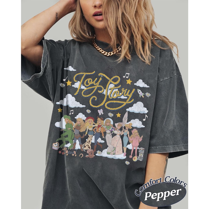 May include: A charcoal grey oversized t-shirt featuring a graphic print with the text Toy Story in yellow rope-style lettering. The design includes illustrations of various characters, white clouds, yellow stars, and musical notes on the front.