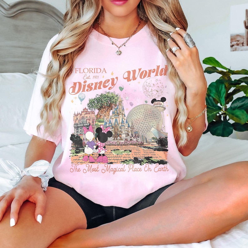 May include: A light pink t-shirt featuring a graphic of Disney World in Florida. The design includes the text "Florida Disney World Est. 1971" and the phrase "The Most Magical Place On Earth." The graphic showcases iconic park elements and characters.