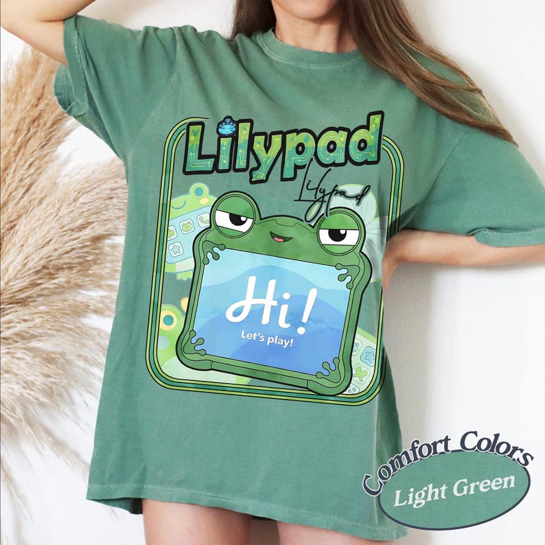 May include: A sage green short-sleeve t-shirt featuring a graphic of a green frog holding a tablet. The tablet screen displays the text "Hi! Let's play!" and the word "Lilypad" is printed above the frog in a stylised font with a green border.