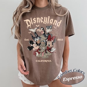 May include: A brown short-sleeve t-shirt featuring a graphic print of Disney characters dressed in Halloween costumes. The text reads Disneyland, Est 1955, and California. The characters include Mickey Mouse, Minnie Mouse, Donald Duck, and Goofy.