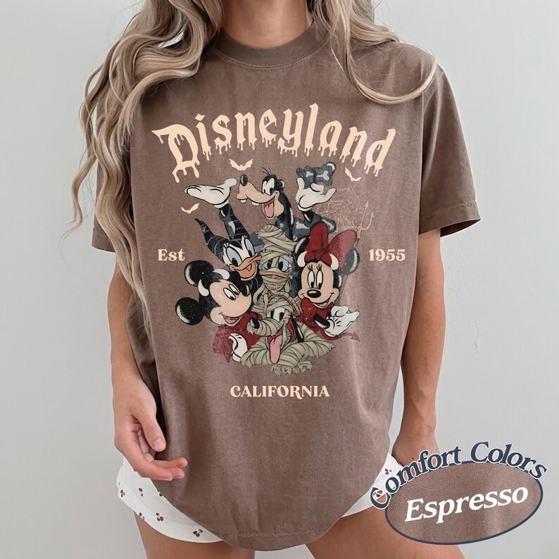 May include: A brown short-sleeve t-shirt featuring a graphic print of Disney characters dressed in Halloween costumes. The text reads Disneyland, Est 1955, and California. The characters include Mickey Mouse, Minnie Mouse, Donald Duck, and Goofy.