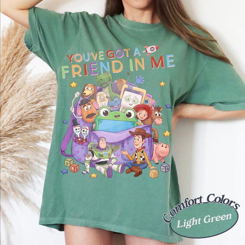 May include: A light green t-shirt featuring a colorful graphic of various animated toy characters and a purple backpack. The text "YOU'VE GOT A FRIEND IN ME" is printed in multicolored letters above the characters. The shirt has short sleeves.