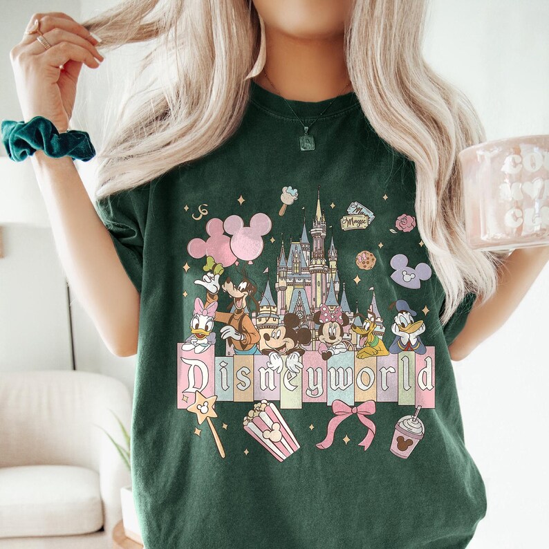 May include: Dark green t-shirt featuring a colorful Disney World graphic. The design includes Mickey and Minnie Mouse, Donald Duck, and a castle. The word "Disneyworld" is displayed in pastel colors.