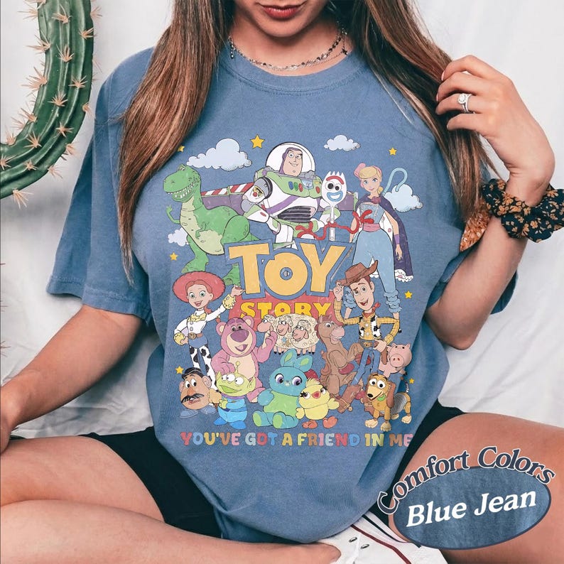 May include: A blue jean colored t-shirt featuring a graphic print of Toy Story characters including Buzz Lightyear, Woody, Jessie, Rex, and others. The text Toy Story and You've Got A Friend In Me are printed in colorful letters on the front of the shirt.