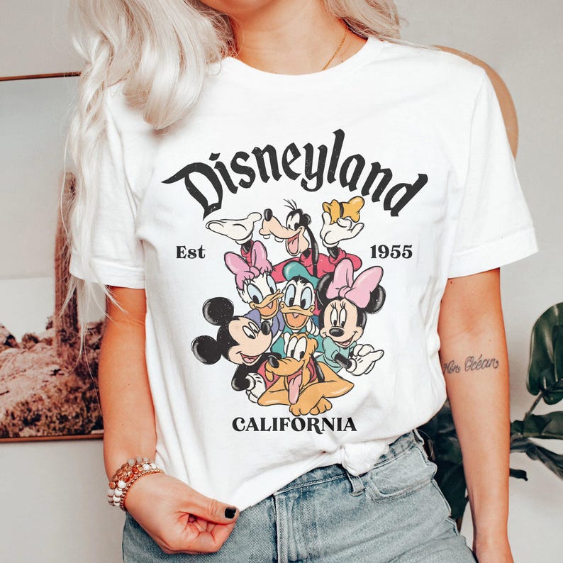 May include: White t-shirt featuring a graphic of Disneyland characters, including Mickey and Minnie Mouse, Donald and Daisy Duck, and Goofy. The text "Disneyland California Est 1955" is printed above the characters in black.