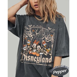 May include: Dark gray t-shirt featuring a Halloween-themed Disneyland graphic. The design includes Mickey Mouse, Minnie Mouse, Donald Duck, and Goofy as skeletons in front of a haunted house. The text "Disneyland" and "Est. 1955" are also visible.