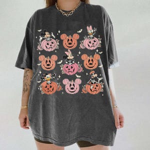 May include: Dark gray t-shirt featuring a Halloween-themed design. The design includes cartoon characters in pumpkins, with bats and floral accents. The pumpkins are in shades of orange and pink.