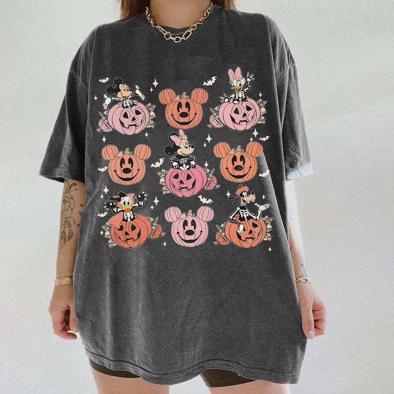 May include: Dark gray t-shirt featuring a Halloween-themed design. The design includes cartoon characters in pumpkins, with bats and floral accents. The pumpkins are in shades of orange and pink.