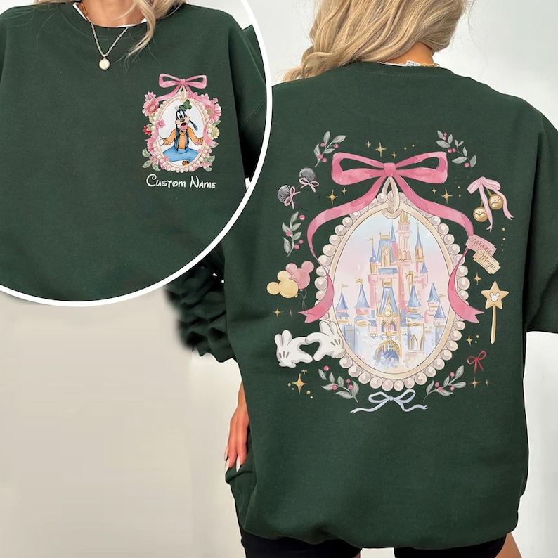 Pode incluir: Camisola verde escuro com um desenho de desenho animado do Pateta em uma moldura floral na frente. A parte de tr&aacute;s da camisola tem um castelo dentro de uma moldura em forma de la&ccedil;o rosa, rodeado por elementos florais e estrelas.