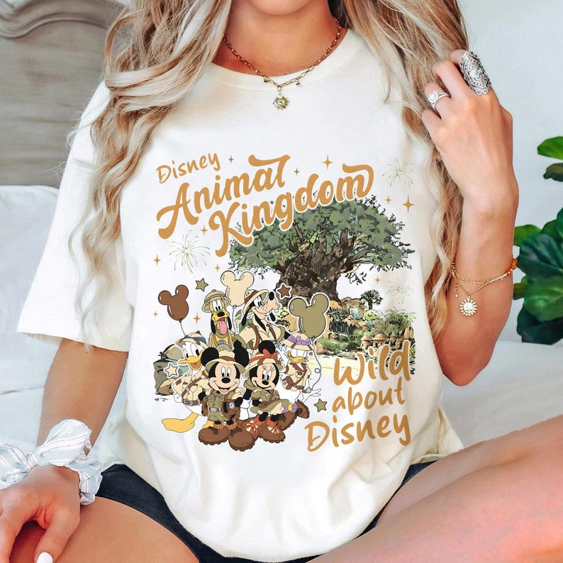 Pu&ograve; includere: T-shirt color crema con la scritta "Disney Animal Kingdom" e "Wild about Disney" e una grafica di personaggi Disney. La grafica include Topolino e Minnie, Paperino e Pippo.