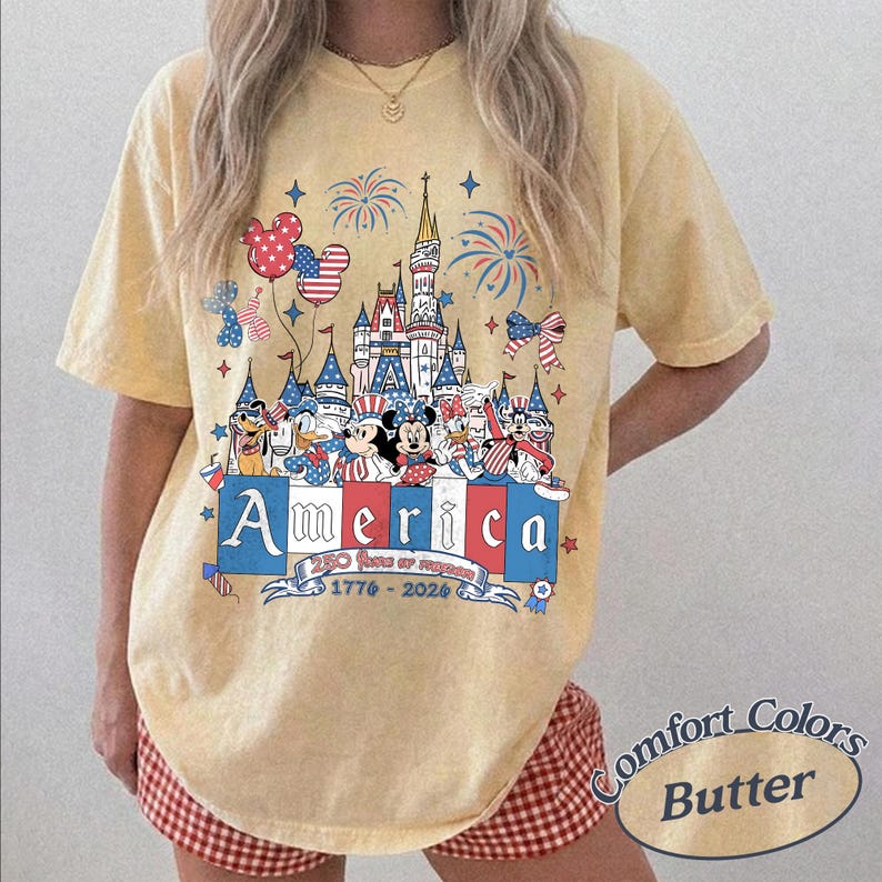 May include: A pale yellow short-sleeve t-shirt featuring a graphic of a castle, fireworks, and cartoon characters in red, white, and blue. The text reads America, 250 years of freedom, and 1776-2026. The design includes star-patterned balloons.