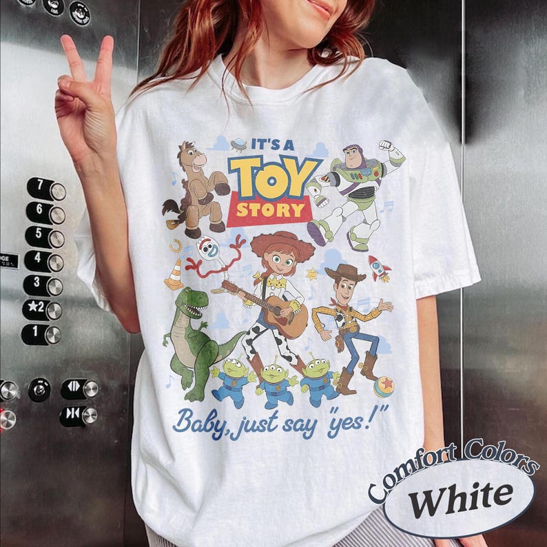 May include: A white t-shirt featuring a colorful graphic print of Toy Story characters including Woody, Jessie, Buzz Lightyear, Rex, Bullseye, Forky, and the Aliens. The text reads IT'S A TOY STORY and Baby, just say yes! in blue and red lettering.