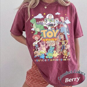 May include: A berry-colored short-sleeve t-shirt featuring a graphic print of various Toy Story characters including Woody, Buzz Lightyear, and Jessie. The text "TOY STORY" and "YOU'VE GOT A FRIEND IN ME" is printed in colorful letters on the front.