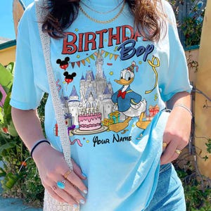 K&ouml;nnte beinhalten: Hellblaues T-Shirt mit einer Grafik von Donald Duck, der Geburtstag feiert. Das Design umfasst ein Schloss, Kuchen, Geschenke und den Text "BIRTHDAY Boy" und "Your Name". Das T-Shirt hat kurze &Auml;rmel.