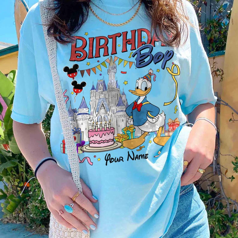 K&ouml;nnte beinhalten: Hellblaues T-Shirt mit einer Grafik von Donald Duck, der Geburtstag feiert. Das Design umfasst ein Schloss, Kuchen, Geschenke und den Text "BIRTHDAY Boy" und "Your Name". Das T-Shirt hat kurze &Auml;rmel.