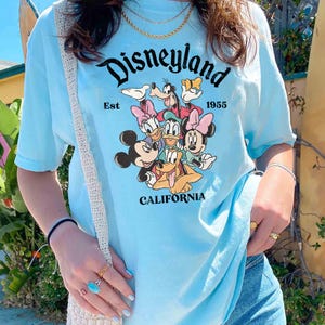 May include: Light blue t-shirt featuring the word "Disneyland" in a script font, with the text "Est 1955" and "California". The shirt displays a graphic of Mickey Mouse, Minnie Mouse, Donald Duck, Daisy Duck, Goofy, and Pluto.