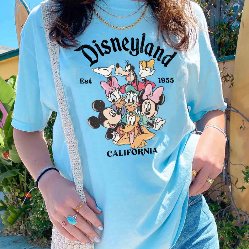 May include: Light blue t-shirt featuring the word "Disneyland" in a script font, with the text "Est 1955" and "California". The shirt displays a graphic of Mickey Mouse, Minnie Mouse, Donald Duck, Daisy Duck, Goofy, and Pluto.