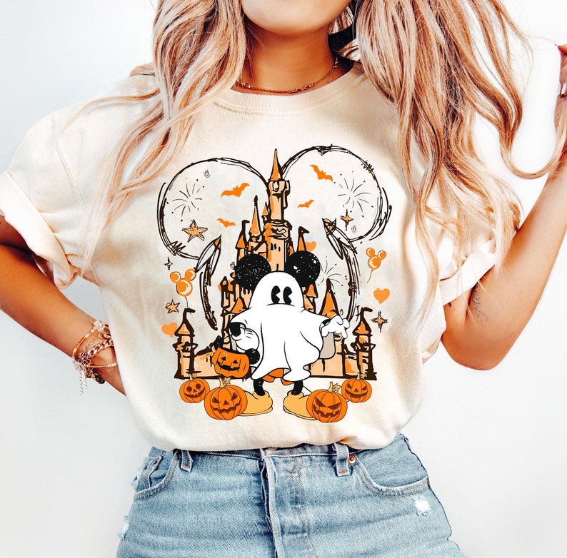 May include: Cream-coloured sweatshirt with a Halloween graphic. The design features a castle, Mickey Mouse ears, a ghost, pumpkins, and fireworks in orange and black. The sweatshirt is a casual, long-sleeved top.