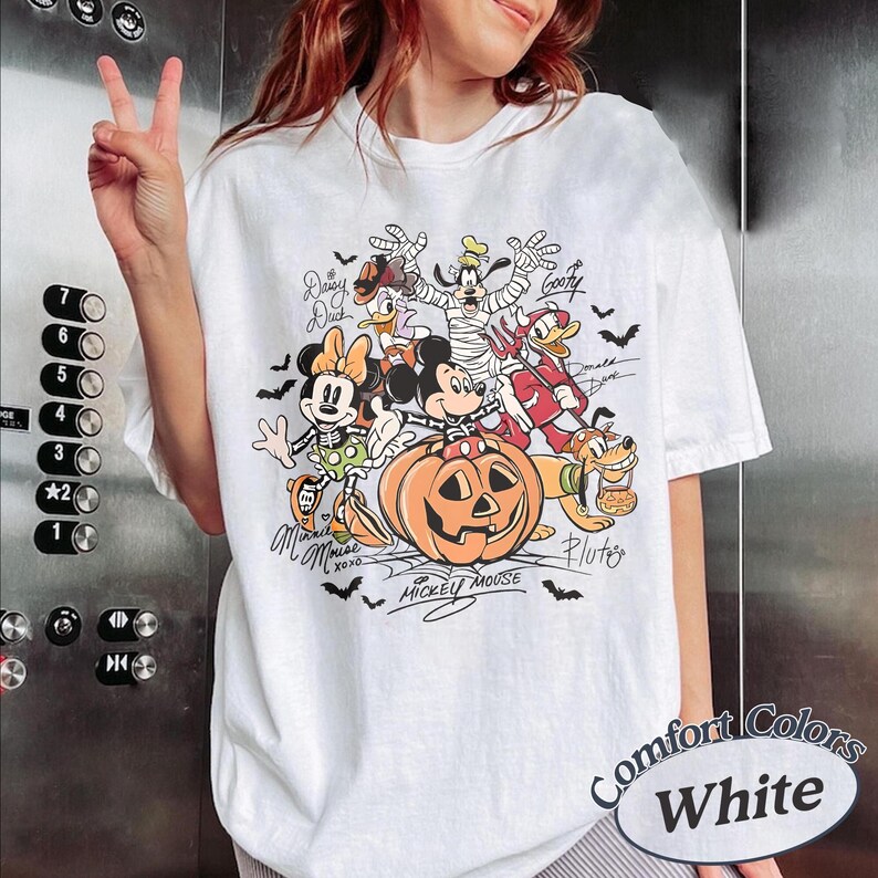 May include: A white t-shirt featuring a colorful Halloween-themed graphic of Disney characters including Mickey Mouse, Minnie Mouse, Donald Duck, Goofy, and Pluto. The characters are dressed in costumes around a large orange jack-o'-lantern pumpkin.