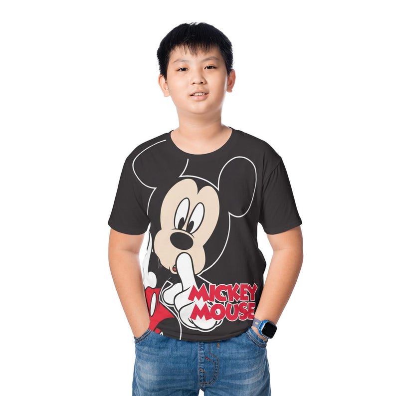 May include: Black t-shirt featuring a large Mickey Mouse graphic. The cartoon character is depicted with a finger to his lips, with the words "Mickey Mouse" in red and white. The shirt is worn by a person wearing blue jeans.