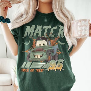 May include: Dark green t-shirt featuring a cartoon image of Mater from the movie Cars. The shirt has the word "MATER" above the image, with "TRICK OR TREAT" and the number "95" below. The shirt is a comfortable, casual style.