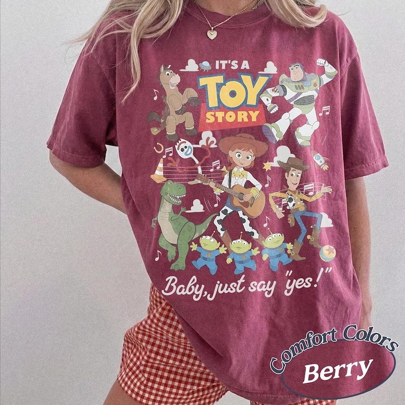May include: A berry-colored short-sleeve t-shirt featuring a graphic print of Toy Story characters including Woody, Buzz Lightyear, Jessie, Rex, Bullseye, Forky, and the Aliens. The text reads IT'S A TOY STORY and Baby, just say yes!