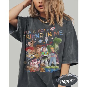 May include: A charcoal gray short-sleeve t-shirt featuring a colorful graphic of various animated characters and the text "YOU'VE GOT A FRIEND IN ME" in a multi-colored font. The shirt has a vintage-style wash and a crew neckline.