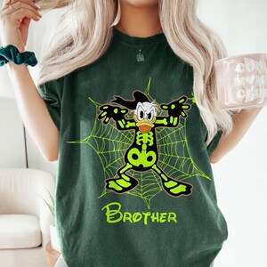 May include: Dark green t-shirt featuring a cartoon character in a skeleton costume, posed in front of a neon green spiderweb. The word "Brother" is printed in neon green at the bottom of the shirt.