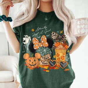 May include: Dark green t-shirt with a Halloween-themed graphic. The design includes a Minnie Mouse head with an orange polka dot bow, a jack-o'-lantern, and the text "Literally Just A Girl". Other elements include a ghost, spiderwebs, and fall leaves.