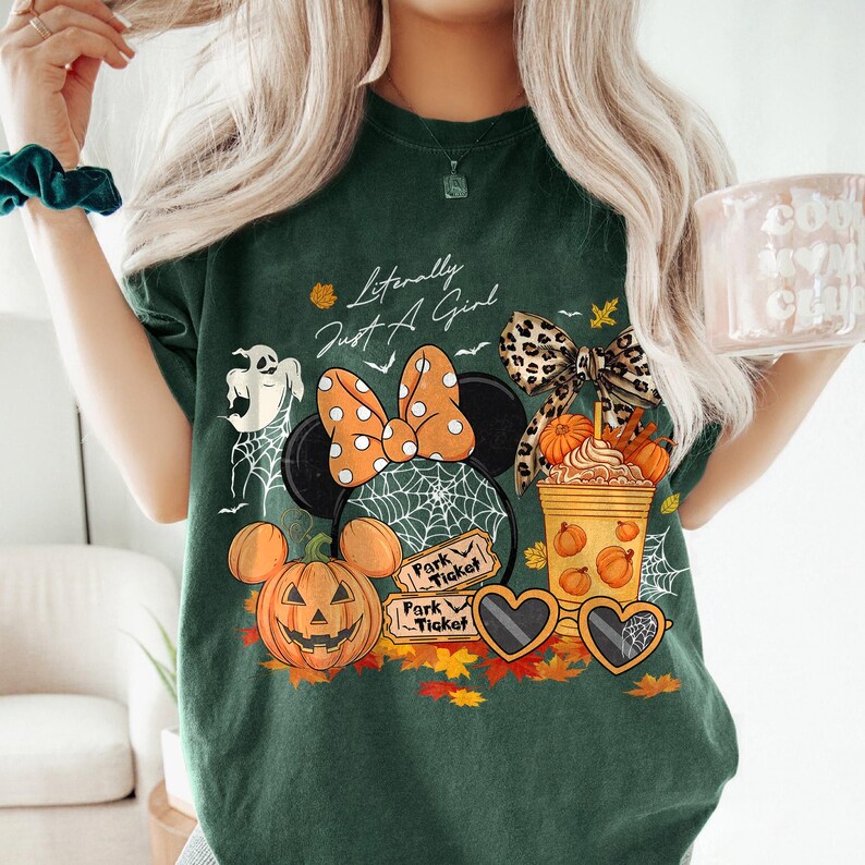 May include: Dark green t-shirt with a Halloween-themed graphic. The design includes a Minnie Mouse head with an orange polka dot bow, a jack-o'-lantern, and the text "Literally Just A Girl". Other elements include a ghost, spiderwebs, and fall leaves.