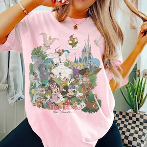 K&ouml;nnte beinhalten: Ein hellrosa T-Shirt mit einem farbenfrohen Aufdruck verschiedener Disney-Figuren, darunter Mickey Mouse, Minnie Mouse und Peter Pan. Der Text "Magic Kingdom" und "Walt Disney World" ist darunter gedruckt. Das T-Shirt hat kurze &Auml;rmel.