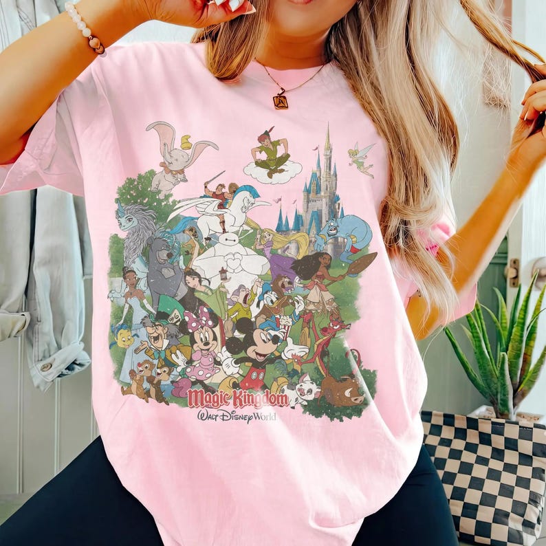 K&ouml;nnte beinhalten: Ein hellrosa T-Shirt mit einem farbenfrohen Aufdruck verschiedener Disney-Figuren, darunter Mickey Mouse, Minnie Mouse und Peter Pan. Der Text "Magic Kingdom" und "Walt Disney World" ist darunter gedruckt. Das T-Shirt hat kurze &Auml;rmel.