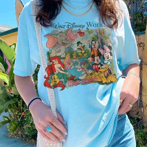 Pode incluir: Camiseta azul clara com uma estampa colorida de personagens da Disney, incluindo Dumbo, Ariel, Mickey Mouse e Minnie Mouse. A camiseta tem o texto "Walt Disney World" e "Magic Kingdom" impresso. A camiseta &eacute; feita de material macio.
