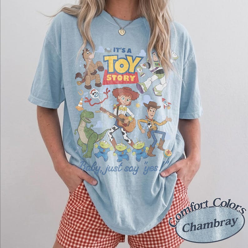 May include: A light blue t-shirt featuring a graphic print of Toy Story characters including Woody, Jessie, Buzz Lightyear, Rex, Bullseye, Forky, and three aliens. The text reads IT'S A TOY STORY and Baby, just say yes. The shirt has a crew neckline.