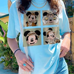 May include: Pale blue t-shirt with a four-panel graphic of Mickey Mouse in safari gear. Each panel depicts Mickey in various poses, wearing a safari hat and khaki clothing, with binoculars in one panel. The background is a muted green.