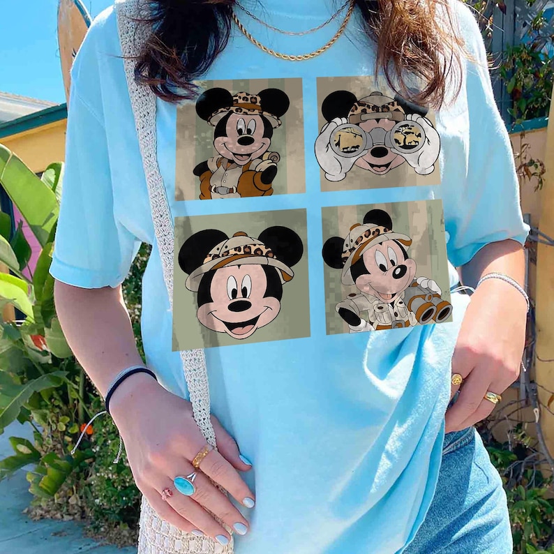 May include: Pale blue t-shirt with a four-panel graphic of Mickey Mouse in safari gear. Each panel depicts Mickey in various poses, wearing a safari hat and khaki clothing, with binoculars in one panel. The background is a muted green.