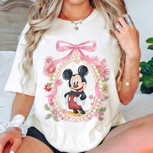 May include: Cream-colored t-shirt featuring a cartoon Mickey Mouse design. Mickey is framed within an oval adorned with pink flowers, a pink bow, and pearls. The design has a vintage aesthetic.