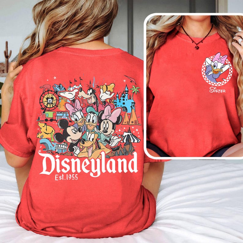May include: Red t-shirt with a Disneyland graphic on the back, featuring cartoon characters and the text "Disneyland Est. 1955". The front displays a Daisy Duck graphic with the word "Sister".