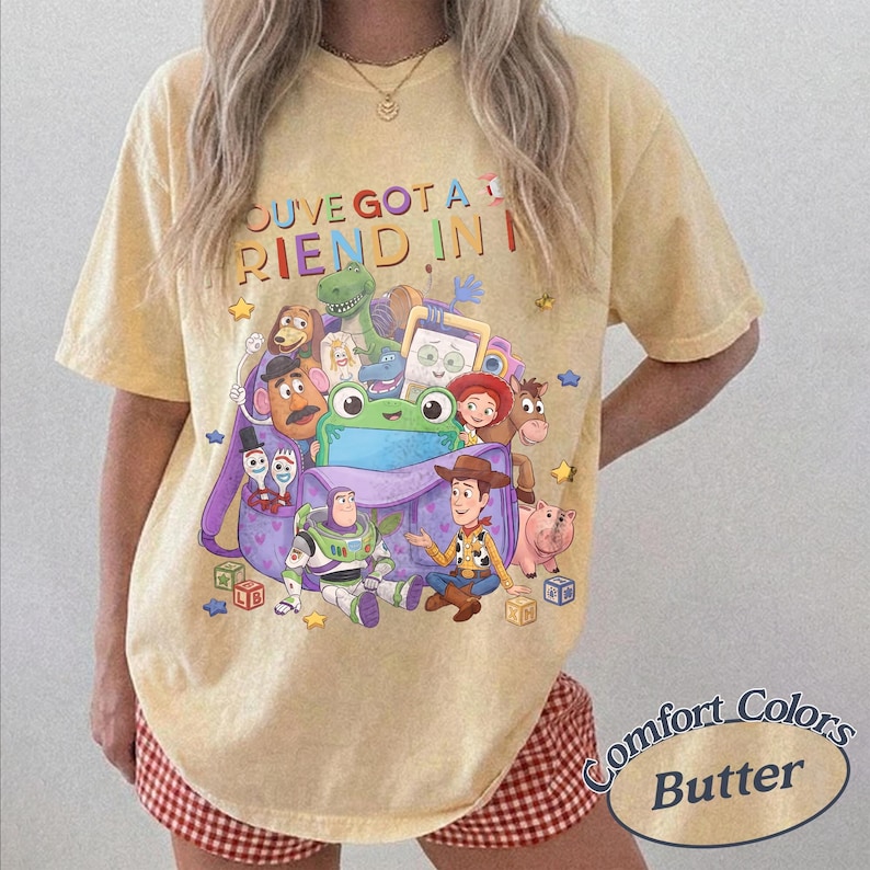 May include: A pale yellow short-sleeve t-shirt featuring a graphic print of various animated characters from Toy Story emerging from a purple backpack. The text "YOU'VE GOT A FRIEND IN ME" is printed in colorful letters above the character illustration.