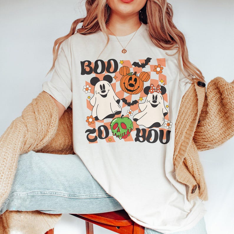 May include: Cream-colored t-shirt with a Halloween-themed graphic. The design features ghosts, a pumpkin, and the text "Boo to You" in a retro font. The shirt has a checkered background with orange and white flowers.