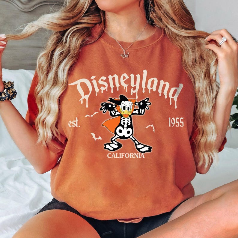 K&ouml;nnte beinhalten: Orangefarbenes T-Shirt mit Disneyland-Grafik. Die Grafik zeigt einen Skelett-Donald-Duck mit Umhang, Flederm&auml;usen und dem Text "Disneyland est. 1955 California."