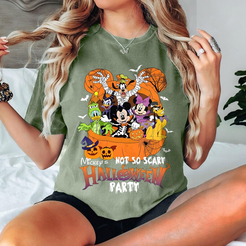 May include: Olive green t-shirt featuring a Halloween-themed graphic. The design includes Mickey Mouse, Minnie Mouse, Donald Duck, Goofy, and other characters dressed in costumes. The text "Mickey's Not So Scary Halloween Party" is also visible.