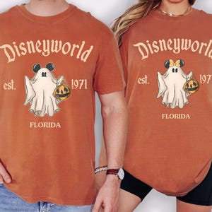 May include: Rust-colored t-shirts with a Disneyworld Halloween design. The graphic features a ghost holding a jack-o'-lantern, with the text "Disneyworld est. 1971 FLORIDA". One shirt has Mickey Mouse ears, the other has Minnie Mouse ears.