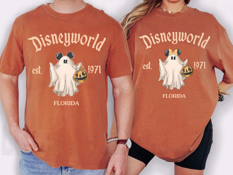 May include: Rust-colored t-shirts with a Disneyworld Halloween design. The graphic features a ghost holding a jack-o'-lantern, with the text "Disneyworld est. 1971 FLORIDA". One shirt has Mickey Mouse ears, the other has Minnie Mouse ears.