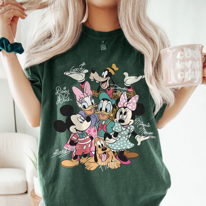May include: Dark green t-shirt featuring a colorful graphic of Disney characters including Mickey Mouse, Minnie Mouse, Donald Duck, Daisy Duck, Goofy, and Pluto. The shirt has the characters' names written above them.