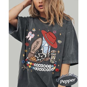 May include: A charcoal grey t-shirt featuring a graphic print of a cow-print cowboy boot with a red hat, a bow, blocks with the letters T, S, and 13, and a beaded necklace. The text reads TAYLOR and I KNEW IT I KNEW YOU. The shirt is labelled Comfort Colors Pepper.
