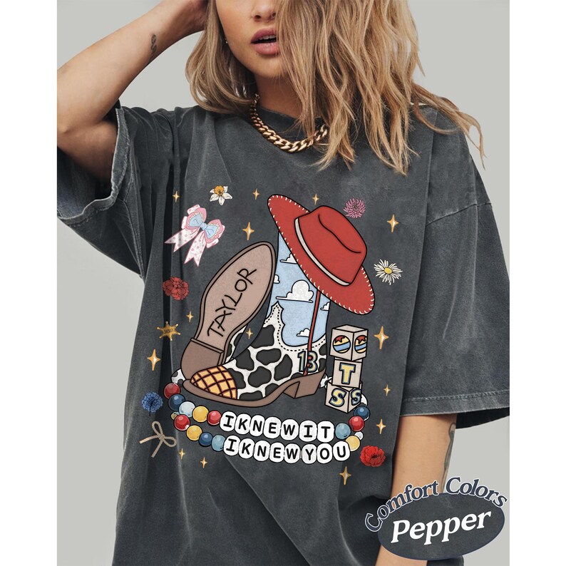 May include: A charcoal grey t-shirt featuring a graphic print of a cow-print cowboy boot with a red hat, a bow, blocks with the letters T, S, and 13, and a beaded necklace. The text reads TAYLOR and I KNEW IT I KNEW YOU. The shirt is labelled Comfort Colors Pepper.
