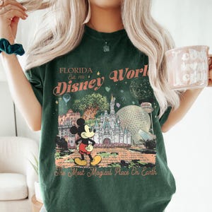 May include: Dark green t-shirt featuring a colorful graphic of Mickey Mouse in front of a Disney World scene. The design includes the text "Florida Disney World" and "The Most Magical Place On Earth."